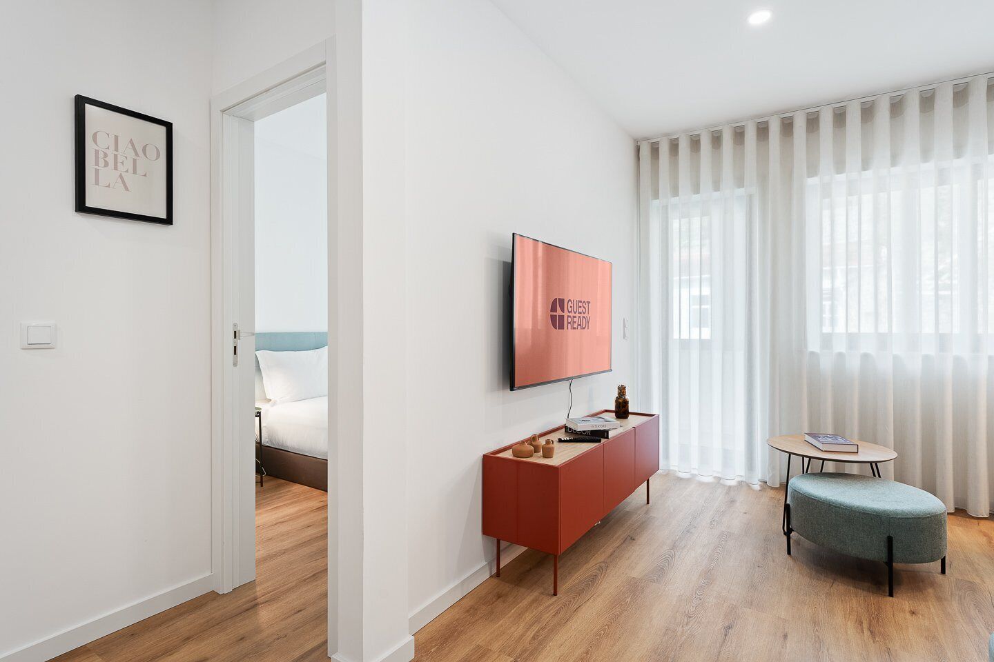 Una camera d'albergo moderna con pavimento in legno, un letto con biancheria bianca, un tavolino consolle rosso con una TV che mostra 'GUEST READY', un piccolo tavolino rotondo con un libro e un quadro incorniciato appeso al muro.