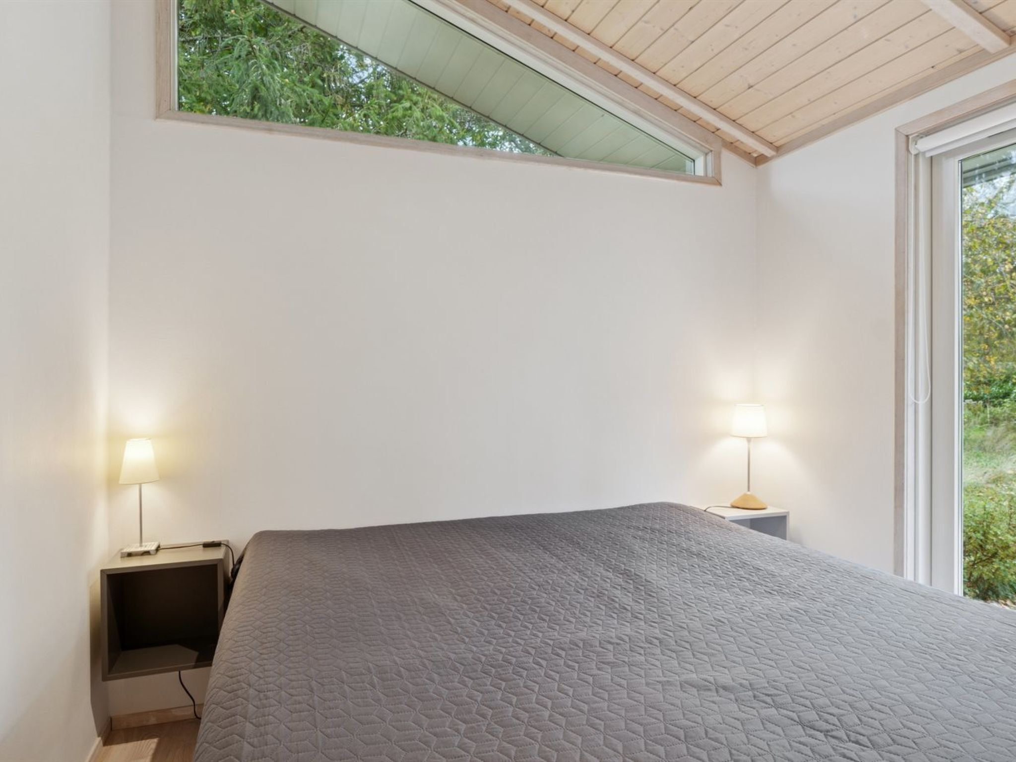 Un dormitorio con una cama grande, dos mesitas de noche con lámparas, un tragaluz y vistas al verdor exterior.