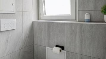 Un baño con inodoro, toallero, ventana y una pequeña repisa con una planta.