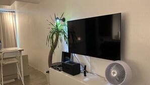 Un salon avec un téléviseur à écran plat fixé au mur, un meuble blanc contenant une platine vinyle et des haut-parleurs, un fauteuil blanc, une petite plante en pot et un ventilateur sur pied, également blanc.