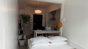 Une petite pièce propre, avec un lit, une table, une plante en pot et un coin cuisine visible à travers une porte ouverte.