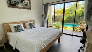 A bedroom with a large bed, a wooden headboard, a desk, and a view of a swimming pool through large sliding doors. - D Krisna Guest House (Canggu)