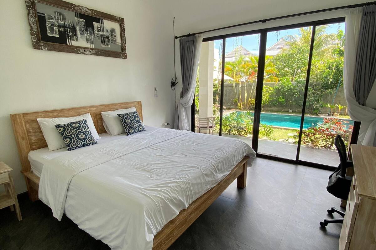 Une chambre dotée d’un grand lit, d’une tête de lit en bois, d’un bureau et offrant une vue sur une piscine grâce à de grandes portes coulissantes.