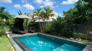 A rectangular swimming pool with a stone edge, surrounded by a well-maintained lawn and various tropical plants. - D Krisna Guest House (Canggu)
