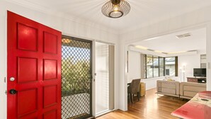 A spacious living area with a red door, a sliding glass door leading to a balcony, a dining area with a table and chairs, and a seating area with a sofa and a TV. - Designed Stay-Convenient, Coastal, Comfortable (Karrinyup)