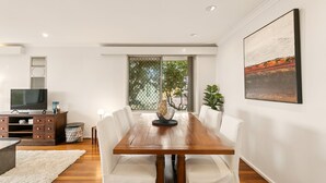 A modern dining area with a wooden table, white upholstered chairs, and a large window with a view of greenery. - Designed Stay-Convenient, Coastal, Comfortable (Karrinyup)