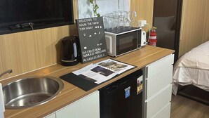 A compact kitchenette with a sink, a coffee maker, a microwave, and a small refrigerator. There is a sign with the address and contact information, and a small shelf with decorative items. - Kalm The Farm 1 by Tiny Away (Numulgi)