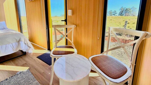 A room with a bed, two chairs, a small table, and a view of a grassy landscape through a large window. - Kalm The Farm 1 by Tiny Away (Numulgi)