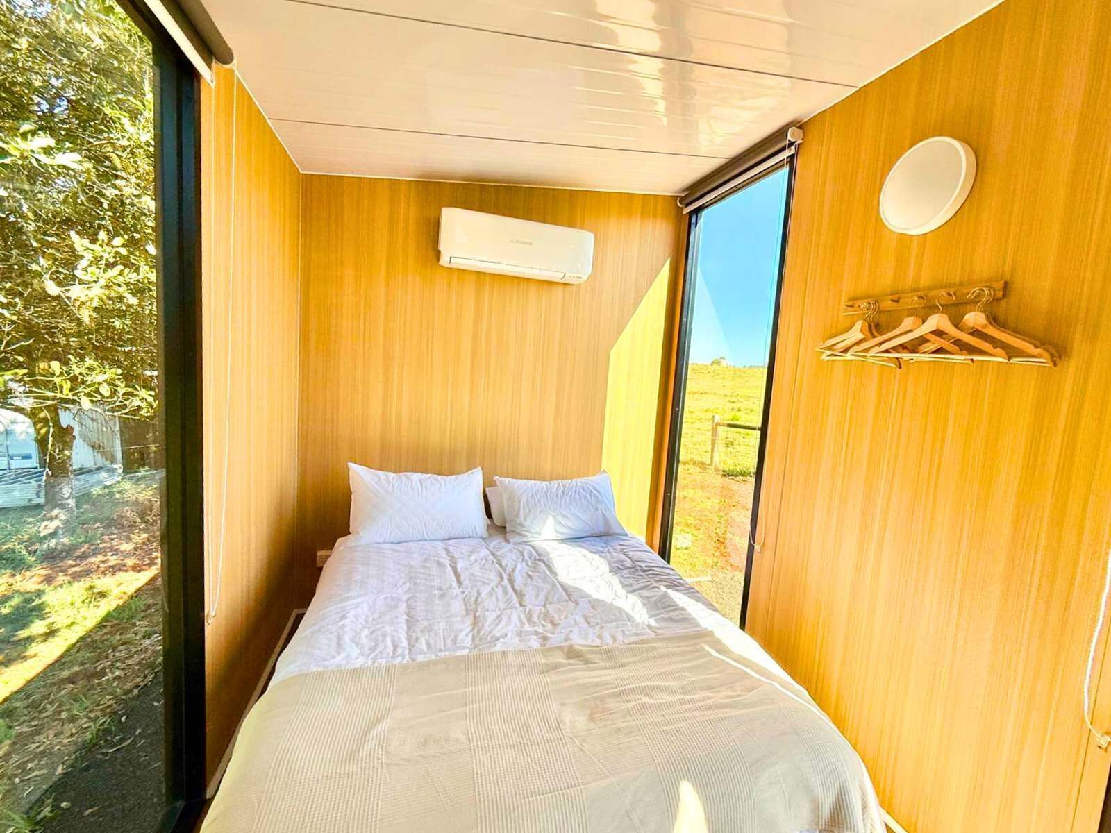 Een slaapkamer met een groot raam, houten wanden, een bed met wit beddengoed en een wandairconditioner.