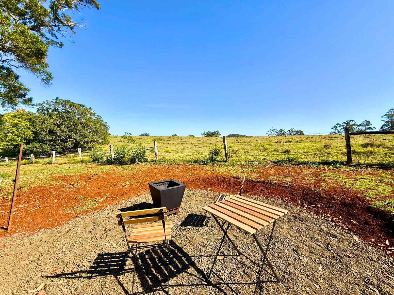 A rustic outdoor seating area with a wooden bench and a metal chair, a black trash bin, and a gravel ground.