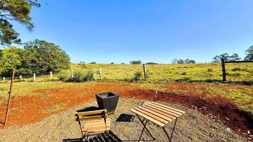 A rustic outdoor seating area with a wooden bench and a metal chair, a black trash bin, and a gravel ground.
