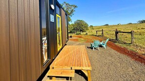 A wooden cabin with a picnic table and chairs outside, a door with a sign, and a fenced area with grass and trees. - Kalm The Farm 1 by Tiny Away (Numulgi)
