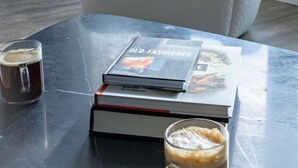 Ein modernes Wohnzimmer mit einem weißen Sessel, einem Couchtisch mit Büchern und einer Kaffeetasse, und Blick auf Bäume durch ein Fenster.