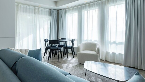 Ein modernes Wohnzimmer mit einem blauen Sofa, einem weißen Sessel, einem runden Couchtisch und einem Essbereich mit Tisch und Stühlen.