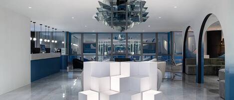 A modern hotel lobby with a central white seating area, a reception desk, and large windows offering views of the cityscape.