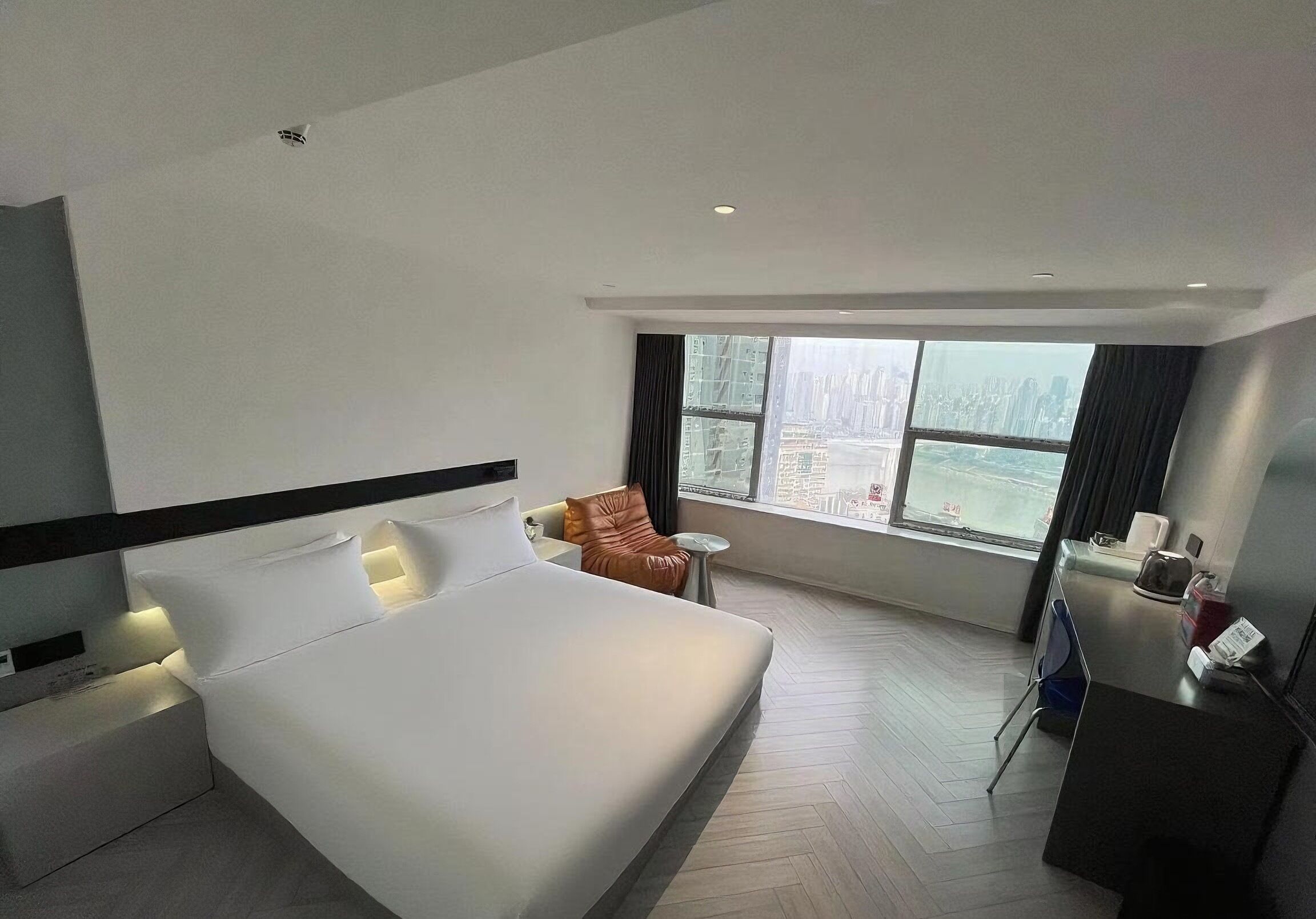 A modern hotel room with a large bed, a small table, a chair, and a view of a cityscape through large windows.