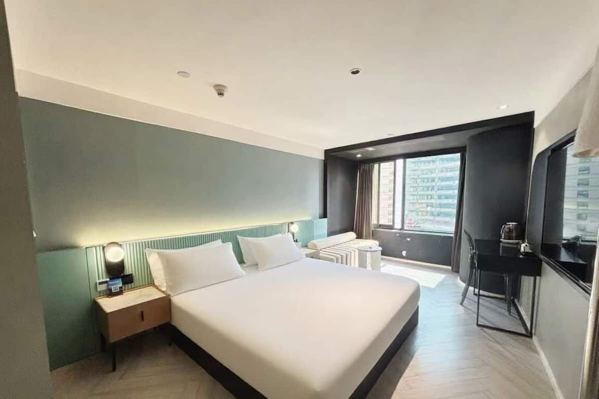 Ein modernes Hotelzimmer mit einem großen Bett, einem Nachttisch mit Lampe, einem kleinen Beistelltisch und Blick auf die Stadt durch ein Fenster.