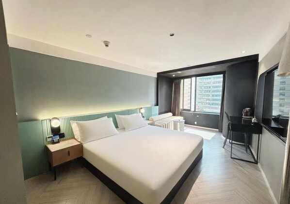 Una habitación de hotel moderna con una cama grande, una mesita de noche con lámpara, una pequeña mesa auxiliar y vistas a la ciudad a través de una ventana.