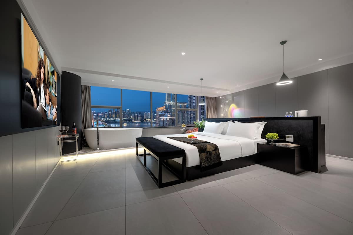 Ein modernes Hotelzimmer mit einem großen Bett, einer Bank, einem Nachttisch und Blick auf die Stadt bei Einbruch der Dunkelheit.