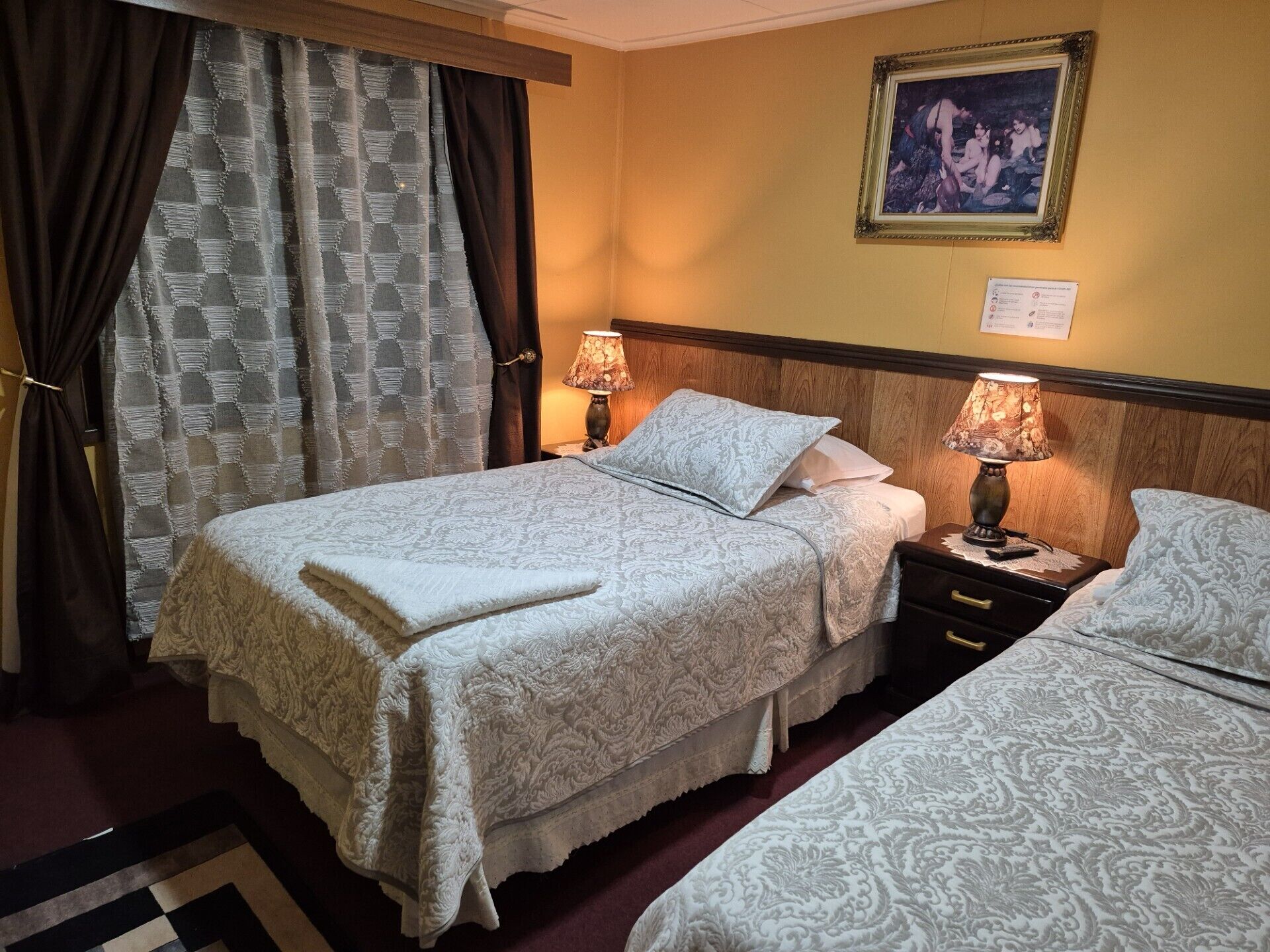 A hotel room with two beds, each with a patterned bedspread and matching pillows. There are two bedside tables with lamps, a wooden headboard, and a framed picture on the wall.