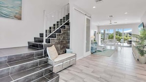 Ein modernes Interieur mit einer Treppe, die zu einem Balkon führt, einer Sitzgelegenheit mit Kissen und Blick ins Freie durch große Fenster.