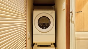 Une petite machine à laver blanche à chargement frontal, installée dans un petit espace buanderie avec une porte en bois et une fenêtre équipée de stores.