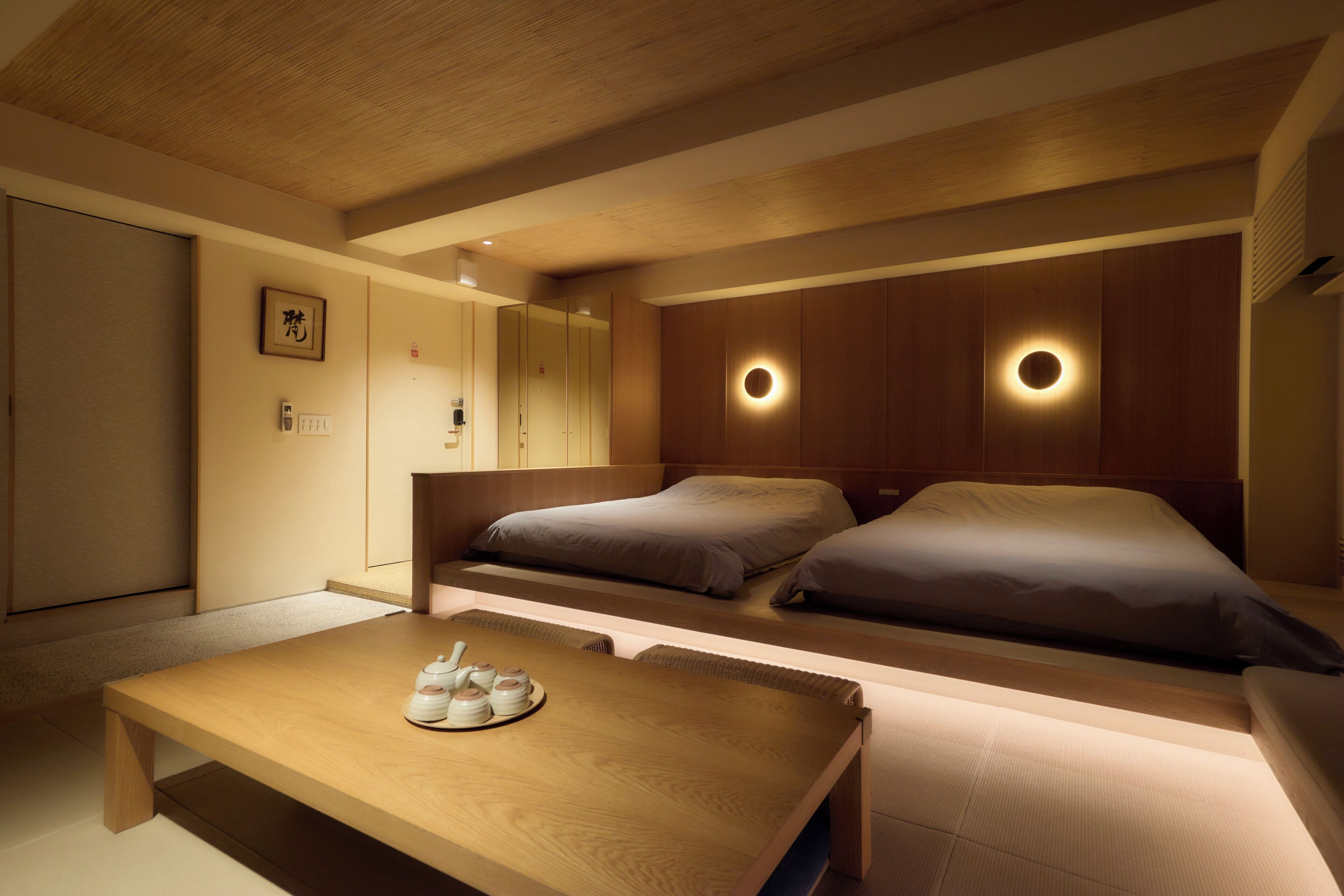 A hotel room with a wooden ceiling, two beds with headboards, a bedside table with a tray of items, and a wall-mounted light fixture.