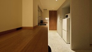 Un couloir avec un plancher en bois, un coin cuisine équipé d’un réfrigérateur et d’un four à micro-ondes, et une porte en bois au bout.