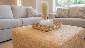 Uma sala de estar com dois sofás, um banquinho de fibra natural e um vaso com uma planta alta.