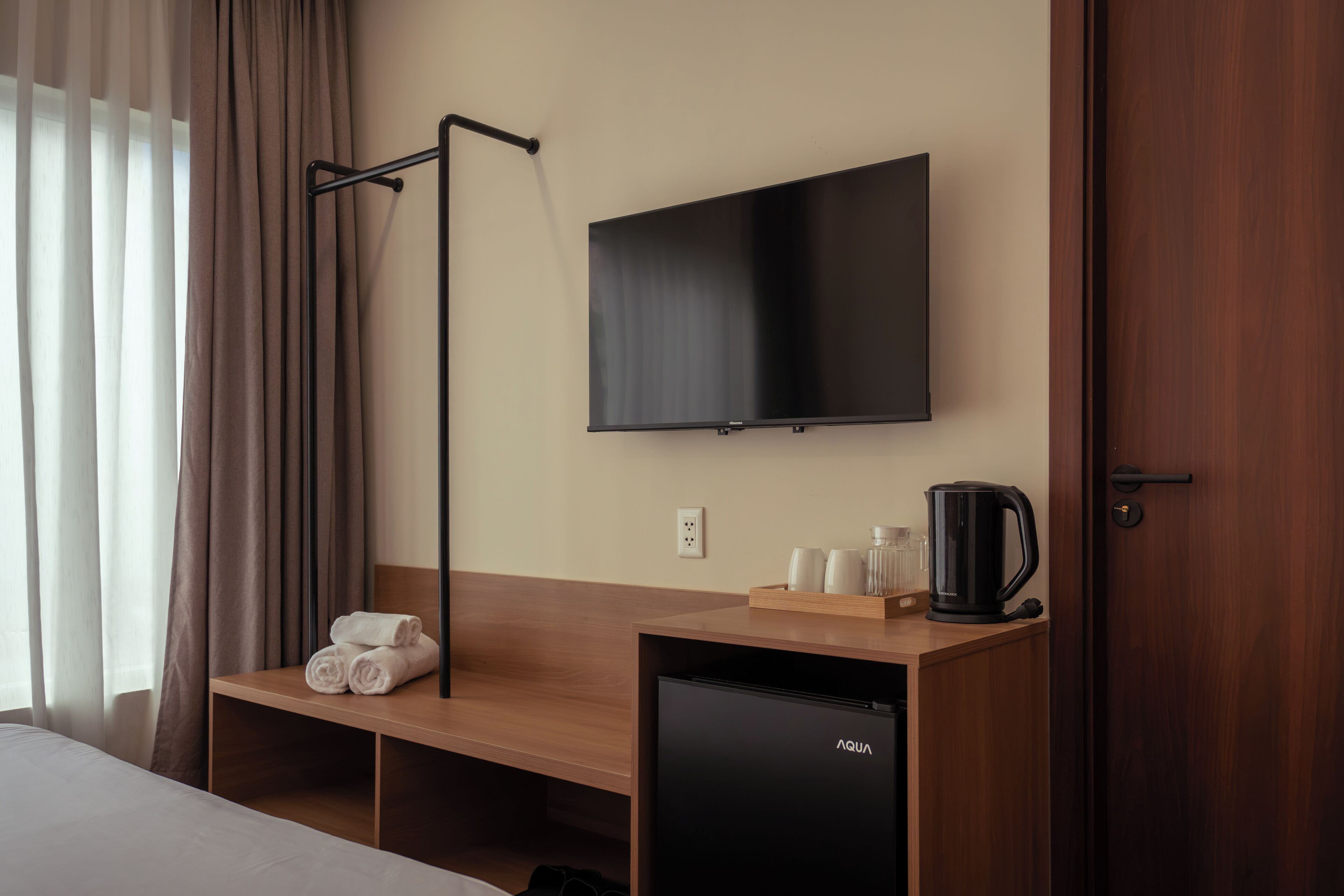 A hotel room with a bed, a TV mounted on the wall, a small refrigerator, a bedside table with a kettle, cups, and a tray, and a wooden bench.