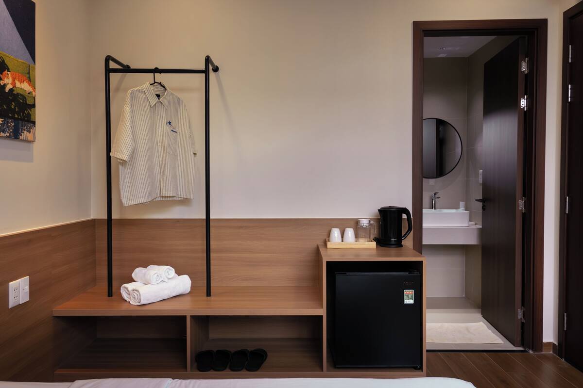 Ein Hotelzimmer mit einem Bett, einem kleinen schwarzen Kühlschrank, einer Holzbk, auf der Handtücher liegen, einem schwarzen Kleiderhaken, einem Waschbecken und einem Spiegel.