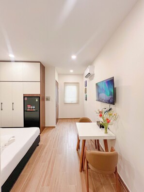 A compact hotel room with a bed, a small table with a chair, a TV mounted on the wall, and a refrigerator.