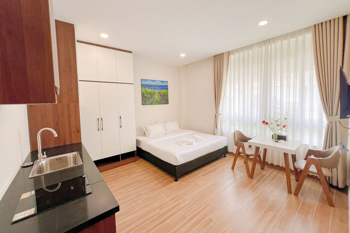 Ein Hotelzimmer mit Bett, Schreibtisch, Stuhl, Fernseher, einem Gemälde und einer Küchenzeile mit Spüle und Herd.