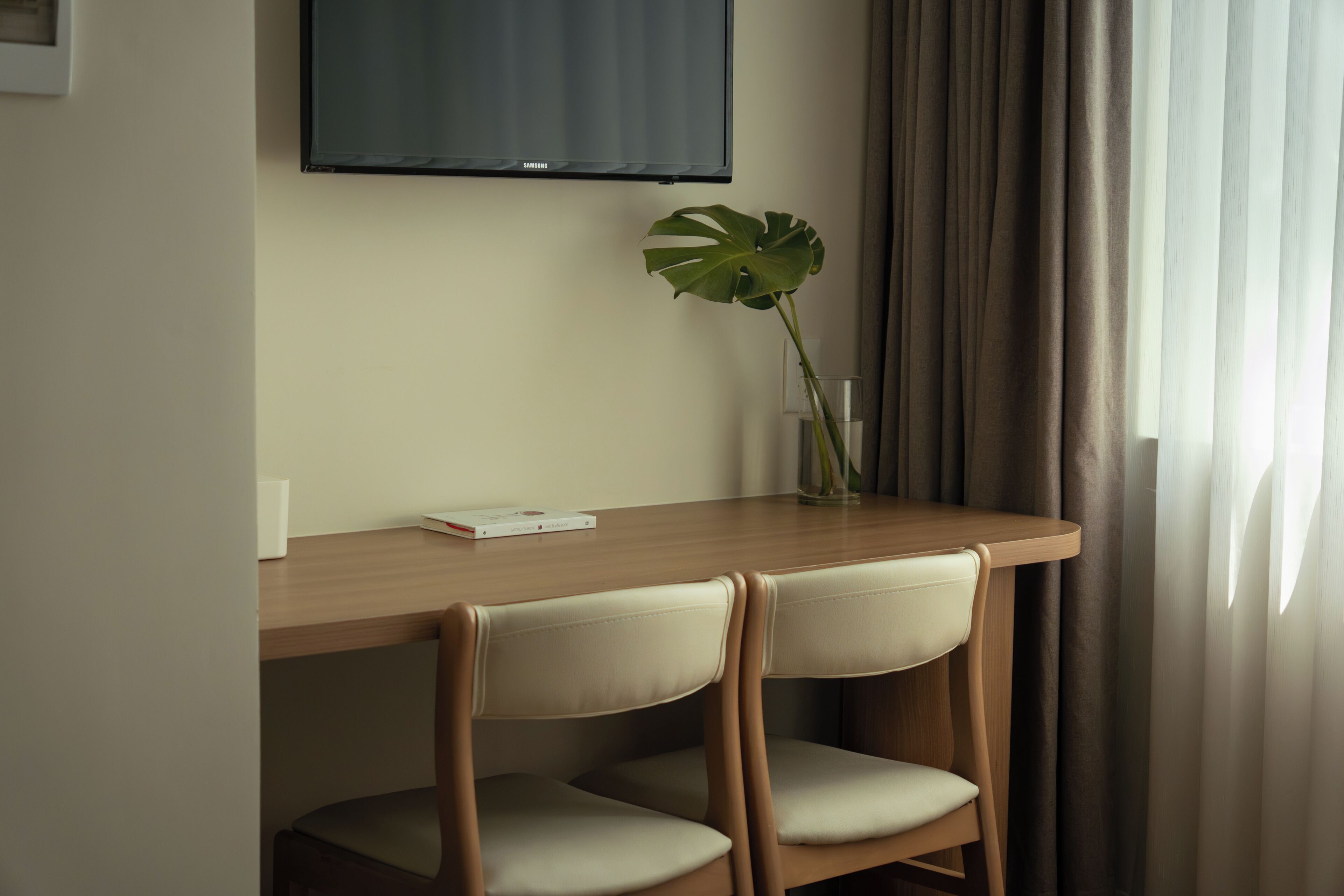 A room with a wooden desk, two chairs, a television mounted on the wall, a plant in a vase, and a window with curtains.