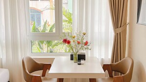 Una stanza con un tavolo bianco, due sedie, un vaso con fiori, una finestra con tende e vista su del verde all'esterno.