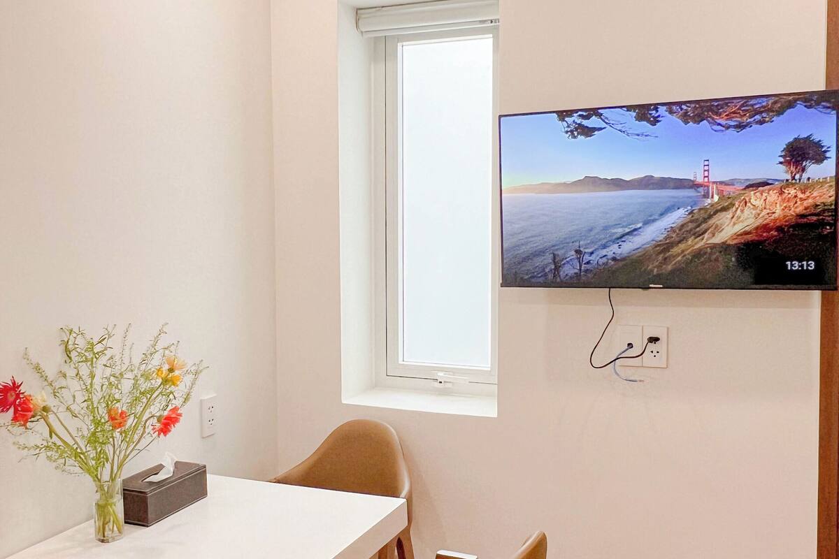 Ein kleiner Essbereich mit Tisch, Stühlen, einer Vase mit Blumen, einem Fernseher an der Wand und einem Fenster.