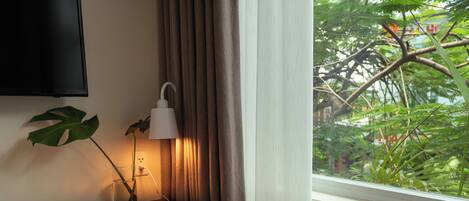 Un rincón de una habitación con un escritorio de madera, una silla, una lámpara, un televisor montado en la pared y una ventana con vistas a zonas verdes.