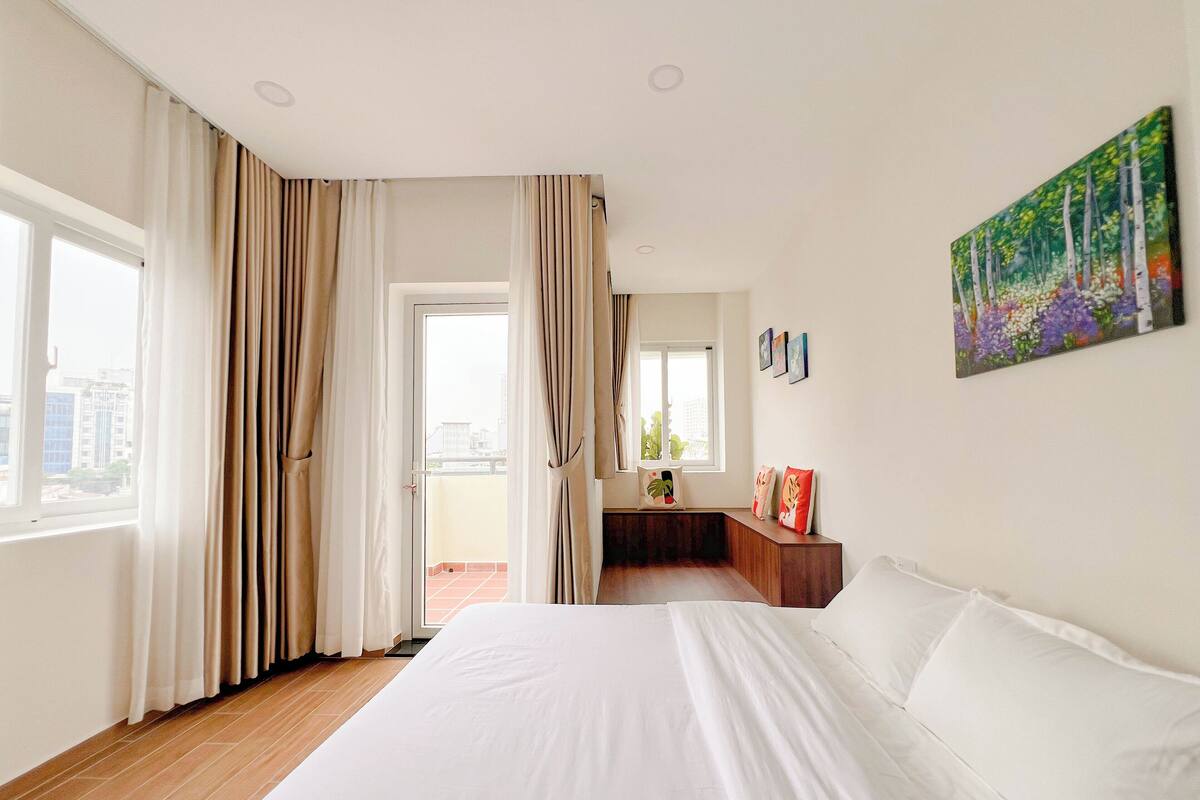Ein Hotelzimmer mit einem großen Bett, einem Balkon mit Vorhängen, einem Gemälde an der Wand und Blick auf die Stadt.