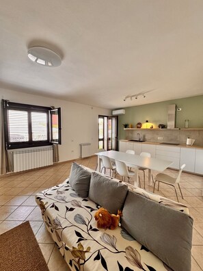 A modern living room with a sofa, a small dog, and a dining area with a kitchen in the background.