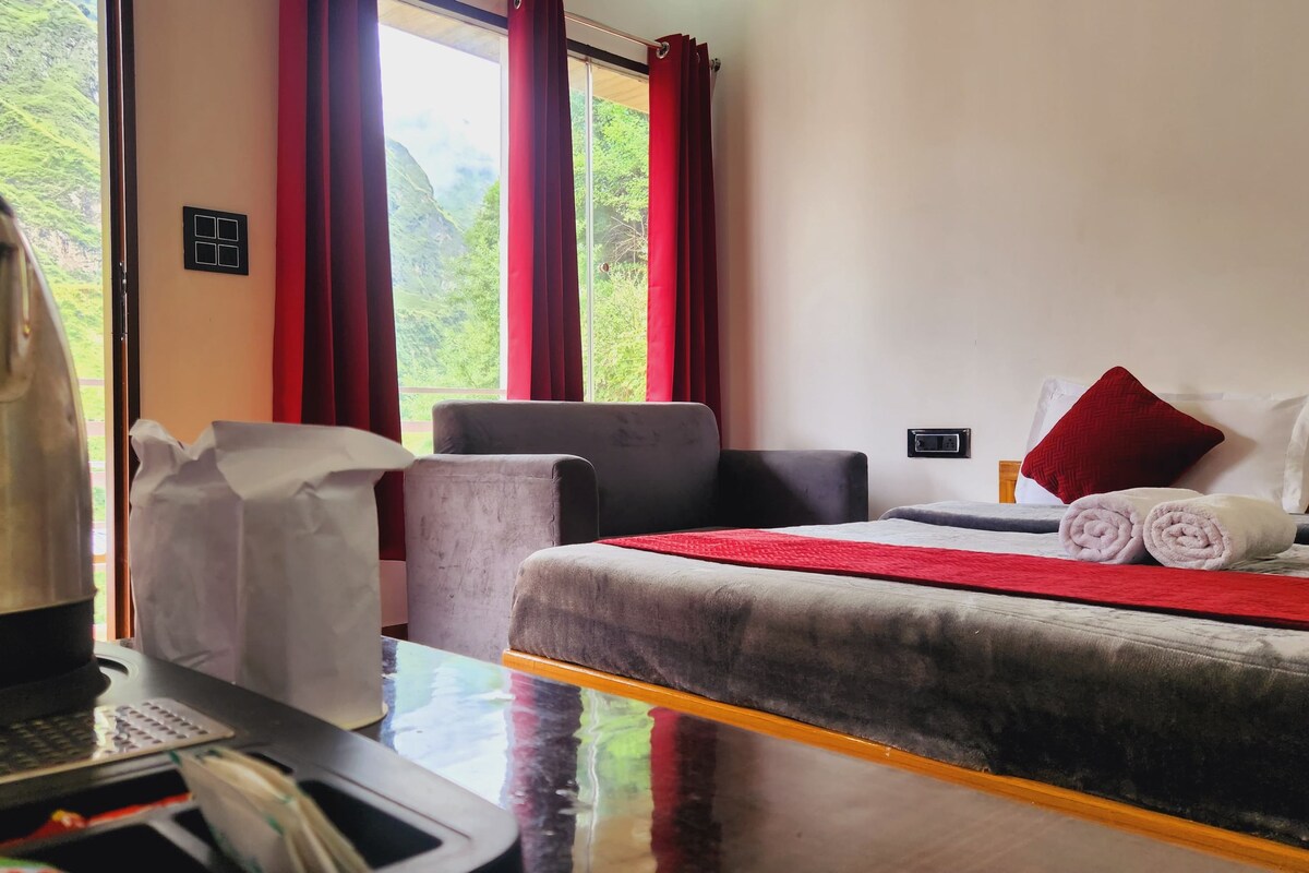 A hotel room with a bed, a desk with a kettle, a chair, and a view of green hills through the window.