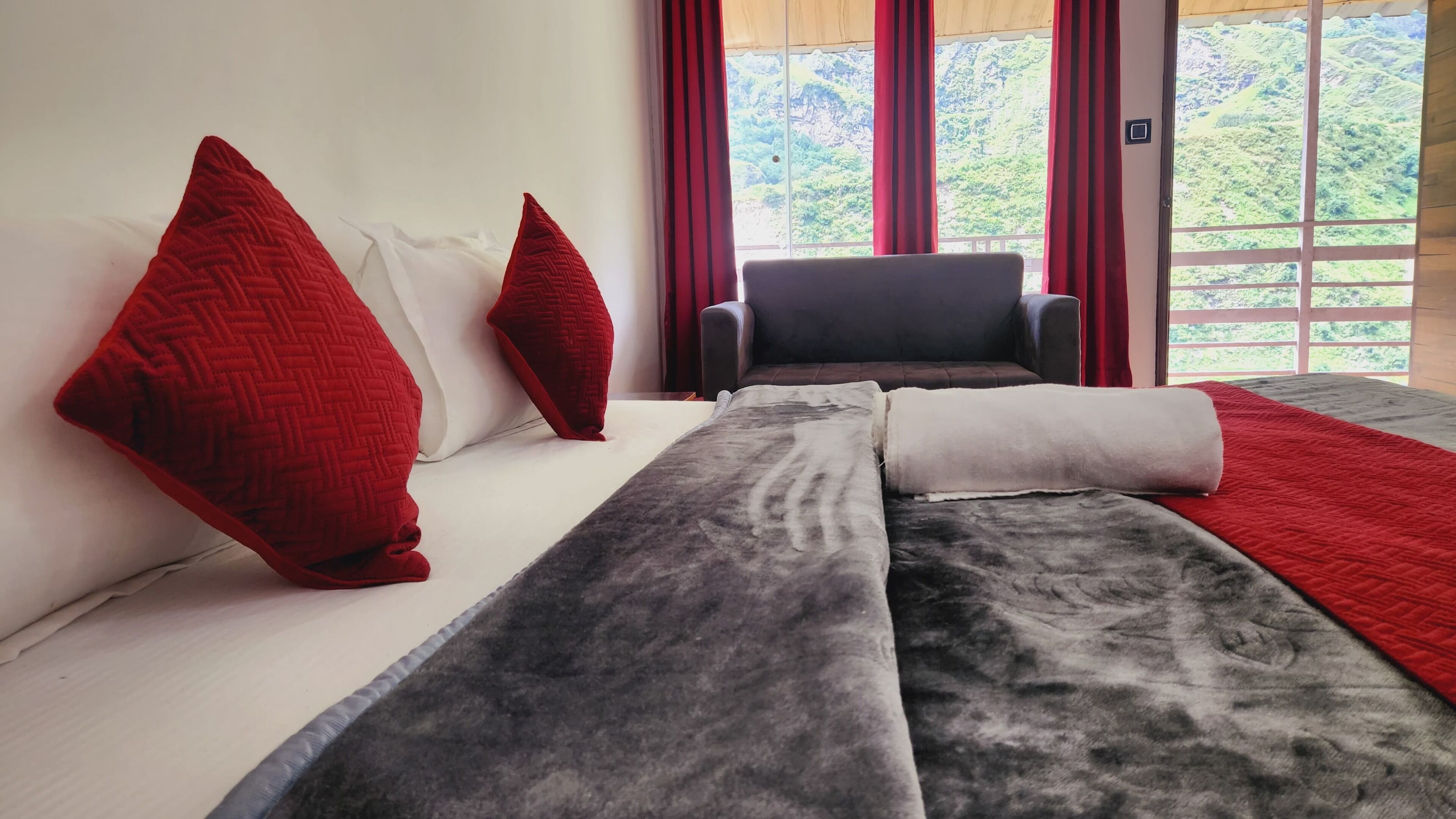 A modern hotel room with a large bed, red pillows, a grey and white blanket, and a view of a mountainous landscape through large windows.