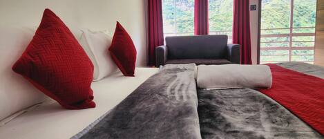A modern hotel room with a large bed, red pillows, a grey and white blanket, and a view of a mountainous landscape through large windows.