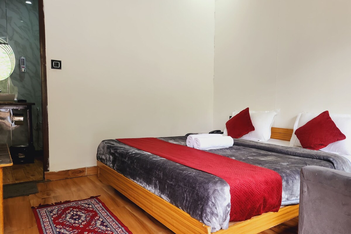 A hotel room with a bed, a wooden nightstand, a red and grey blanket, white pillows, and a red throw.