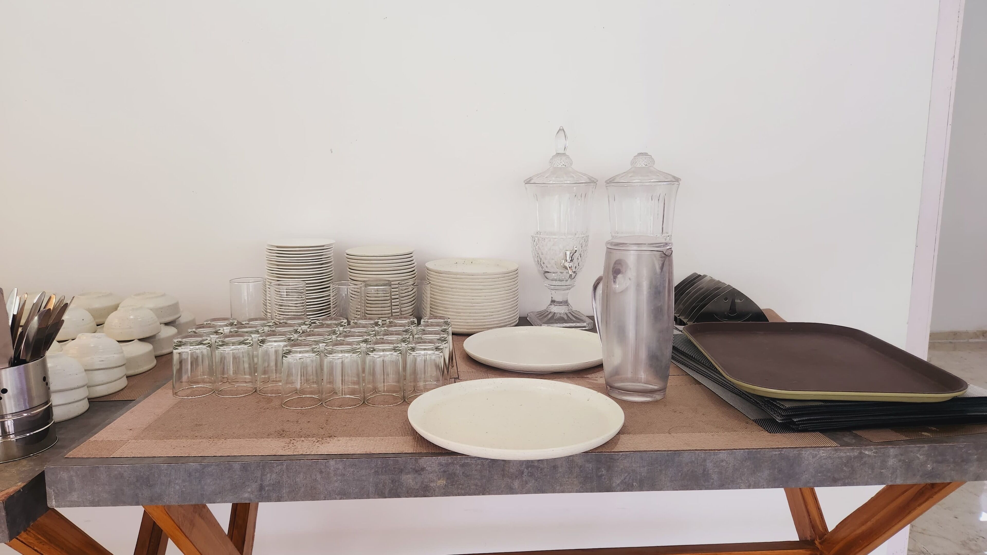 Una mesa con torres de platos blancos, vasos transparentes, una jarra y una bandeja.