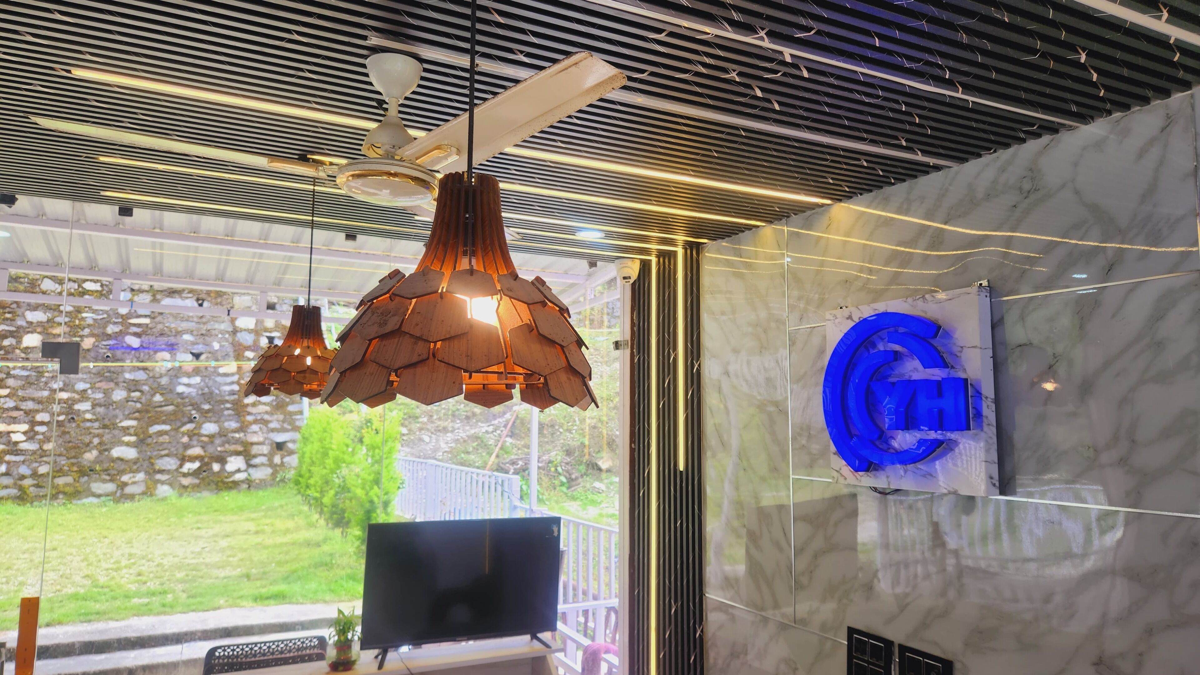 A modern interior with a ceiling fan, wooden pendant lights, a large TV, and a marble wall with a logo.