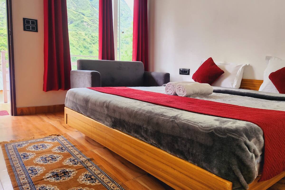 Habitación de hotel con cama de madera, ropa de cama roja y gris, un sillón gris y vistas a colinas verdes a través de amplias ventanas.