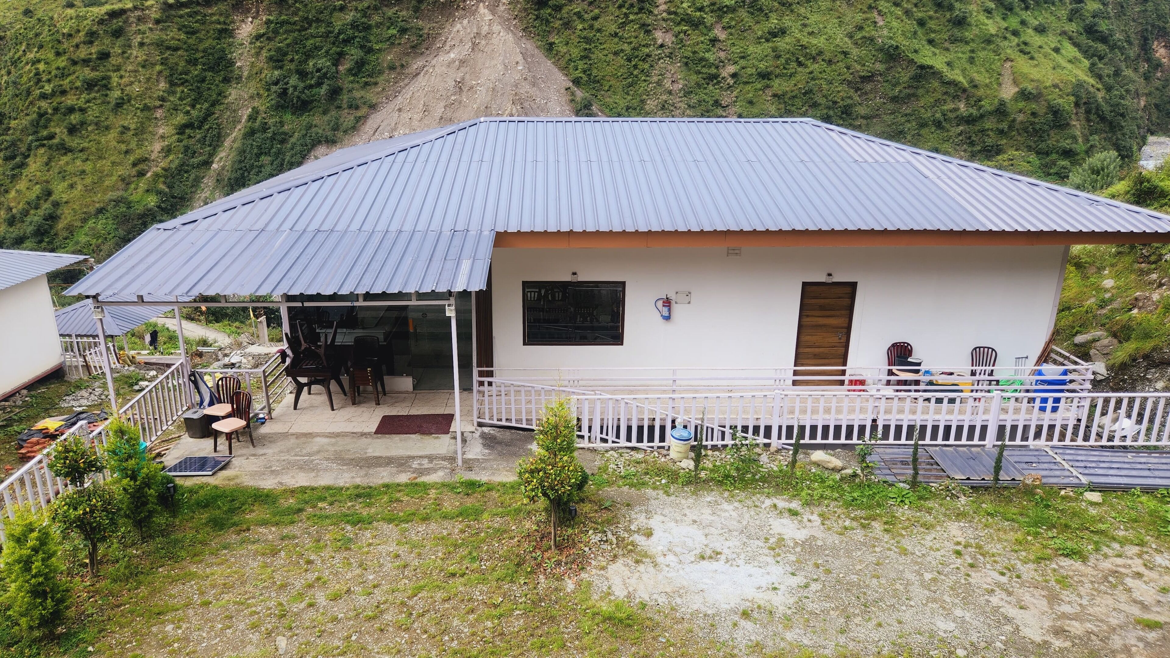 Un edificio pequeño con techo metálico, un patio cubierto con sillas y mesas, y vistas a un paisaje montañoso.