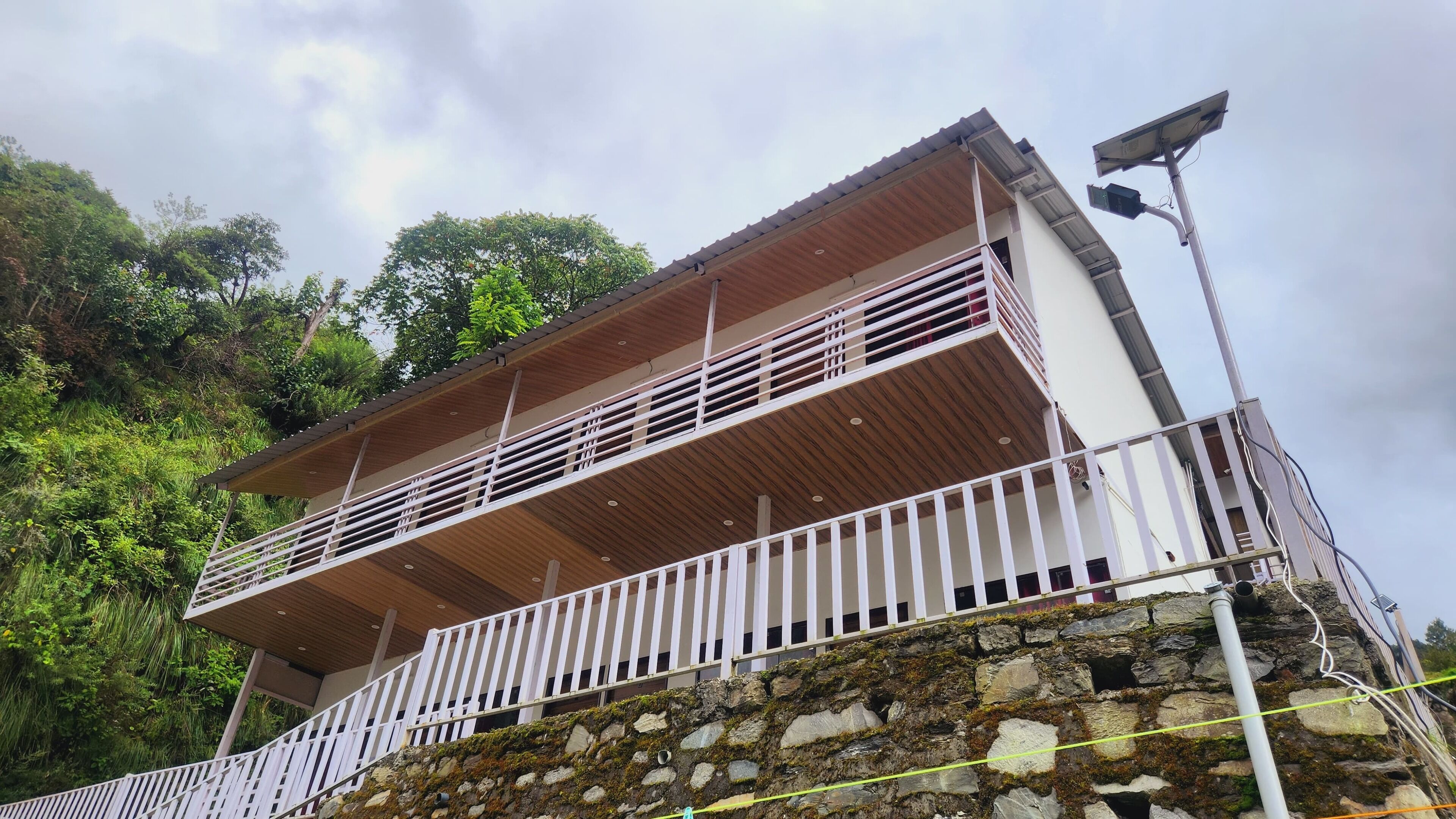 A two-story building with a balcony, surrounded by greenery and a stone wall.