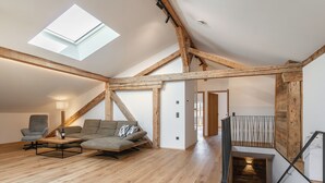 A spacious loft with a high ceiling, exposed wooden beams, a skylight, a cozy seating area with a sofa and armchairs, and a staircase with a rustic design.
