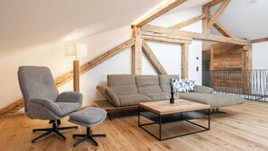 A modern loft with exposed wooden beams, a grey armchair, a sofa, and a wooden coffee table with glasses and a bottle.